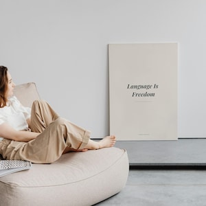 Puede incluir: Un cuadro decorativo rectangular con el texto Language Is Freedom en una fuente serif negra sobre un fondo beige claro. La lámina está colocada en posición vertical sobre un suelo gris junto a un puf beige y una pared blanca.