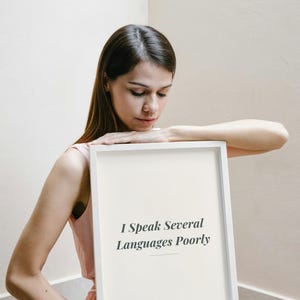 Puede incluir: Un marco rectangular blanco que contiene una impresión de color crema con el texto negro "I Speak Several Languages Poorly" centrado en una fuente serif. El marco tiene un borde limpio y sencillo y se sostiene frente a un fondo neutro.