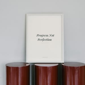 Puede incluir: Una pieza de arte mural rectangular con marco blanco que presenta el texto negro Progress Not Perfection centrado sobre un fondo crema claro. El marco es fino y blanco, y la obra está colocada sobre tres pedestales cilíndricos rojo oscuro.
