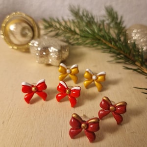 Könnte beinhalten: Nahaufnahme von mehreren schleifenförmigen Ohrsteckern in Rot, Gold und Gelb. Die Ohrringe sind auf einer hellen Holzoberfläche angeordnet, mit Weihnachtsornamenten und Grünzeug im Hintergrund. Die Schleifen haben ein glänzendes Finish.