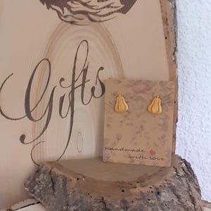 Könnte beinhalten: Goldfarbene Kürbis-Ohrstecker auf einer Karte mit der Aufschrift "handmade with love". Die Ohrringe werden auf einem rustikalen Holzregal mit dem Wort "Gifts" in Schreibschrift präsentiert.