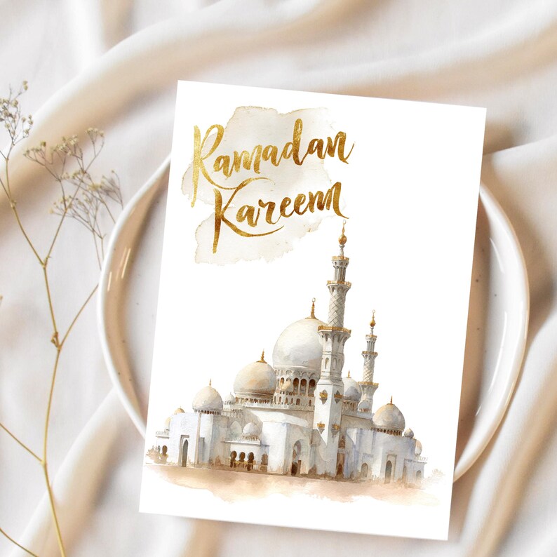 Puede incluir: Una tarjeta de felicitaci&oacute;n blanca con la inscripci&oacute;n dorada "Ramadan Kareem" sobre una imagen de acuarela de una mezquita. La mezquita tiene c&uacute;pulas y minaretes blancos con detalles dorados. La tarjeta est&aacute; sobre un plato blanco.