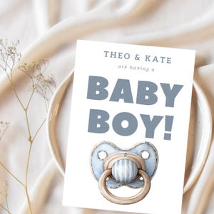 May include: A baby shower invitation card with the text "THEO & KATE are having a BABY BOY!" in blue. The card features a watercolor illustration of a blue and white striped pacifier. The card is placed on a white plate.