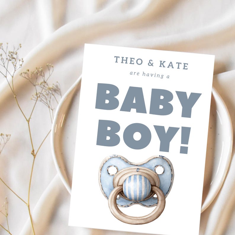May include: A baby shower invitation card with the text "THEO & KATE are having a BABY BOY!" in blue. The card features a watercolor illustration of a blue and white striped pacifier. The card is placed on a white plate.