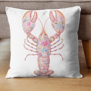 May include: A square throw pillow featuring a white background with a central illustration of a lobster. The lobster is light pink and decorated with a detailed pattern of blue, pink, and white flowers and green leaves across its entire body.