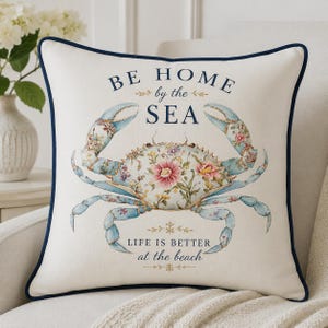 May include: A square off-white throw pillow with a dark blue piped edge. The center features a blue crab illustration decorated with pink and white floral patterns. The text reads BE HOME by the SEA and LIFE IS BETTER at the beach in blue.