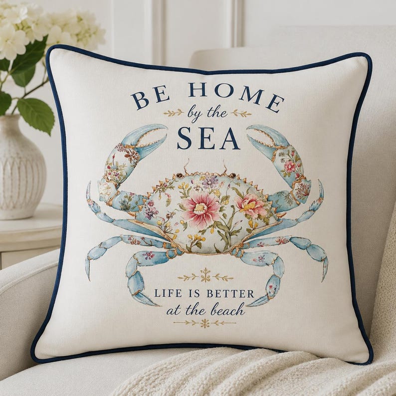 May include: A square off-white throw pillow with a dark blue piped edge. The center features a blue crab illustration decorated with pink and white floral patterns. The text reads BE HOME by the SEA and LIFE IS BETTER at the beach in blue.