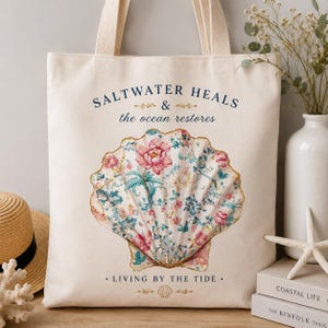 May include: A cream-colored canvas tote bag featuring a large central illustration of a seashell filled with a pink and blue floral pattern. The text reads SALTWATER HEALS & the ocean restores and LIVING BY THE TIDE with small decorative gold accents.