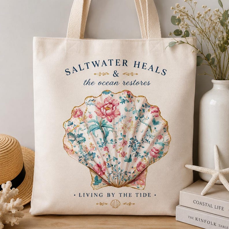 May include: A cream-colored canvas tote bag featuring a large central illustration of a seashell filled with a pink and blue floral pattern. The text reads SALTWATER HEALS & the ocean restores and LIVING BY THE TIDE with small decorative gold accents.