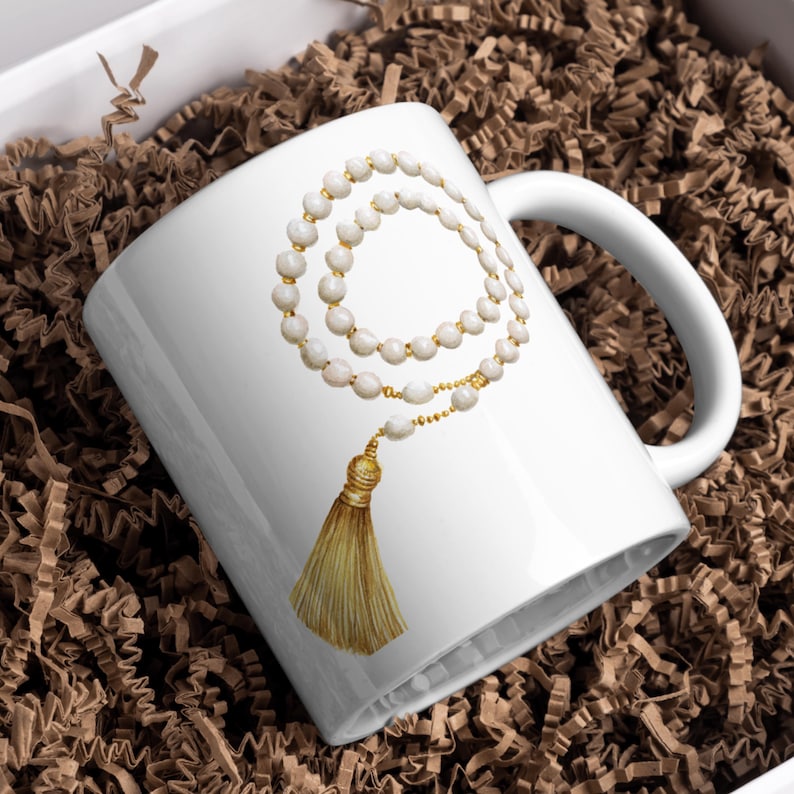 Puede incluir: Taza de cer&aacute;mica blanca con un dise&ntilde;o de acuarela de un collar de cuentas y una borla dorada. El dise&ntilde;o del collar incluye cuentas blancas y detalles dorados. La taza tiene un asa blanca y est&aacute; sobre un fondo de material de embalaje marr&oacute;n.