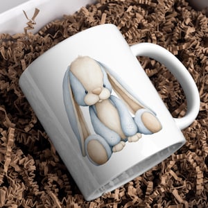 May include: White ceramic mug featuring a watercolor illustration of a blue and beige bunny. The bunny has long ears and is sitting down. The mug is on a bed of brown paper shreds, suggesting it is packaged for gifting.