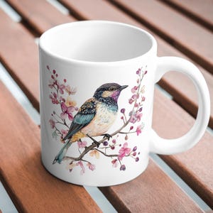 May include: White ceramic mug featuring a watercolor illustration of a colorful bird perched on a branch with pink blossoms. The bird has blue, yellow, and purple feathers. The mug has a curved handle.