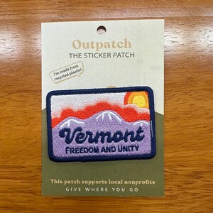 May include: A rectangular embroidered patch on a display card. The patch features a purple mountain range, a red and orange sunset, and the text Vermont FREEDOM AND UNITY. The card reads Outpatch THE STICKER PATCH and I'm made from recycled plastic.