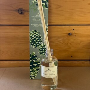 May include: A clear glass reed diffuser bottle filled with liquid and a bundle of wooden reeds, positioned in front of a tall rectangular box featuring a green pinecone pattern and the text Soap & Paper Factory Roland Pine Siberian fir.