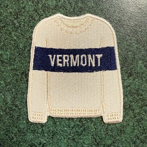 May include: An embroidered patch shaped like a cream-colored long-sleeve sweater with a ribbed crew neckline and hem. A horizontal navy blue stripe runs across the chest, featuring the word VERMONT stitched in white capital letters.