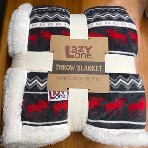 May include: A folded throw blanket with a dark gray background, featuring a repeating pattern of red moose and white zigzag stripes. The blanket has a white sherpa fleece trim and a cardboard label reading Lazy One Throw Blanket 50" by 60".