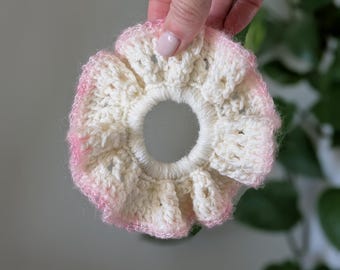 May include: A handmade crochet scrunchie in cream with a pink ombre edge. The scrunchie has a ruffled design and a circular opening in the center. This hair accessory is perfect for adding a touch of handmade charm to any hairstyle.