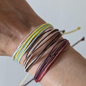 May include: A set of thin, multi-colored string bracelets worn on the wrist. The bracelets feature a variety of colors including pale yellow, light blue, white, dark brown, tan, deep purple, and red, arranged in several layered strands.