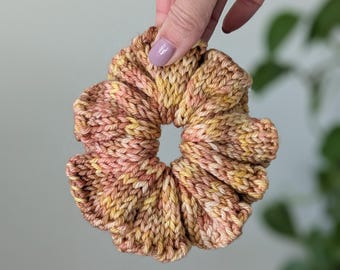 May include: A handmade scrunchie in shades of yellow, peach, and brown. The knitted fabric is gathered to create a voluminous, circular shape. The scrunchie is held up by a hand with painted nails.