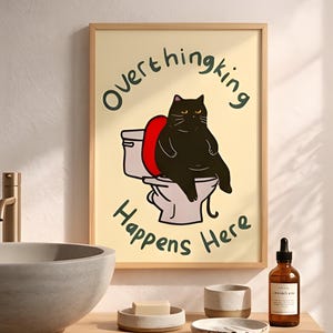 Op de afbeelding: Een ingelijste print met een zwarte kat op een toilet. De tekst luidt "Overthinking Happens Here". De kat heeft gele ogen en zit op een rode bril. De achtergrond is lichtgeel.