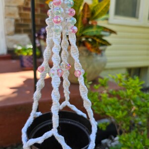 May include: A white macrame plant hanger with pink and blue iridescent beads. The plant holder is designed to hold a small, dark brown pot. The background includes greenery and a house.