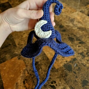 May include: A handmade, dark blue crochet witch hat with a white crescent moon applique. The hat features a curled tip and long, dangling ties. The brim is wide and floppy, and the hat is held by a hand.