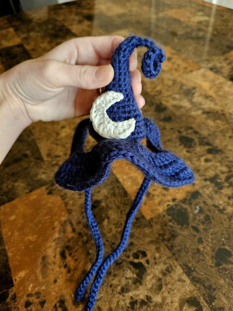 May include: A handmade, dark blue crochet witch hat with a white crescent moon applique. The hat features a curled tip and long, dangling ties. The brim is wide and floppy, and the hat is held by a hand.