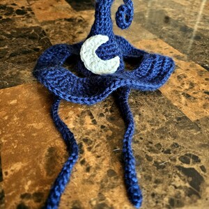 May include: A handmade, dark blue crochet witch hat with a white crescent moon applique. The hat features a pointed top with a spiral curl and two long, braided ties. The hat is displayed on a brown surface.