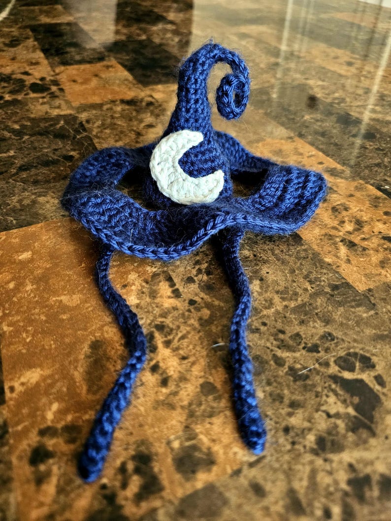 May include: A handmade, dark blue crochet witch hat with a white crescent moon applique. The hat features a pointed top with a spiral curl and two long, braided ties. The hat is displayed on a brown surface.
