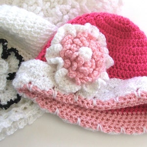 Peut inclure: Un chapeau crochet&eacute; rose et blanc avec une grande fleur blanche sur le dessus. Le chapeau est fait avec une vari&eacute;t&eacute; de points et a une bordure rose et blanche.