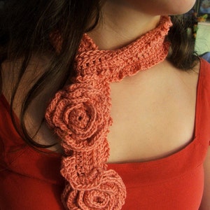 Peut inclure: Une écharpe en crochet corail avec un motif floral. L'écharpe est portée autour du cou d'une personne portant une chemise rouge.