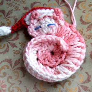 Puede incluir: Un adorno navideño de crochet rosa y blanco con forma de Papá Noel durmiendo. El adorno tiene un gorro rojo con ribete blanco y ojos azules.