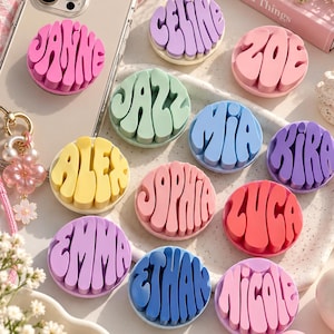 Pode incluir: Uma coleção de suportes circulares para telemóvel com letras em relevo de estilo ondulado em várias cores pastel e vibrantes. Cada suporte apresenta um nome único, incluindo Janine, Celine, Zoe, Jazz, Mia, Kira, Alex, Sophia, Luca, Emma, Ethan e Nicole.