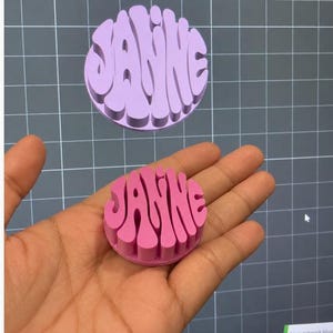 May include: A circular, three-dimensional object featuring the name JANNE in a bold, wavy, raised font. The physical object is bright pink, while the digital rendering displayed on the screen behind it is a lighter shade of lavender.
