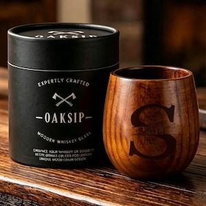 Può includere: Un bicchiere da whisky in legno marrone scuro con una grande lettera S incisa e il nome Scott, accompagnato da una scatola regalo cilindrica nera etichettata OAKSIP con un logo a forma di asce e testo che descrive il bicchiere in legno.