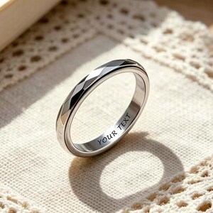 May include: A silver ring with a geometric design and the engraved text "YOUR TEXT". The ring is displayed on a white lace doily, with a book and necklace in the background. The ring's design features multiple facets.