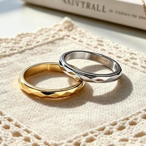 May include: Two rings, one gold and one silver, with geometric facets, sit on a cream lace doily. A gold necklace with a small pendant and a book titled "NAIVTRALL" are in the background.