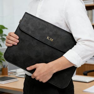 Puede incluir: Funda negra para portátil con cierre de solapa, sostenida por una persona. La funda tiene las iniciales doradas "K.M" en la parte delantera. La persona lleva una camisa blanca abotonada. El fondo incluye un escritorio y una estantería.