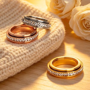 May include: Three rings in gold, rose gold, and silver, each with a band of clear, sparkling stones. The rings are displayed on a cream-colored knitted surface, with soft lighting and a blurred background of white roses.
