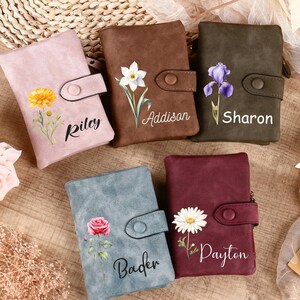 Könnte beinhalten: Fünf personalisierte Geldbörsen in den Farben Rosa, Braun, Grün, Blau und Burgunderrot. Jede Geldbörse hat ein Blumendesign und einen Namen. Die Geldbörsen sind auf einer Holzoberfläche mit floralen Akzenten angeordnet.