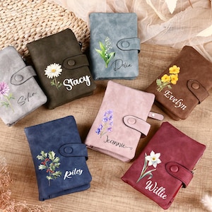 Puede incluir: Una colección de carteras personalizadas en varios colores, incluyendo gris, verde oliva, azul, rosa, marrón y burdeos. Cada cartera presenta un diseño floral y un nombre. Las carteras están dispuestas sobre una superficie de madera.