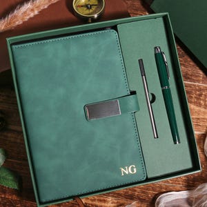 May include: A green leather-bound journal with a silver clasp, a pen, and a stylus, presented in a matching green box. The journal is embossed with the initials "NG". The set is displayed on a wooden surface with decorative elements.
