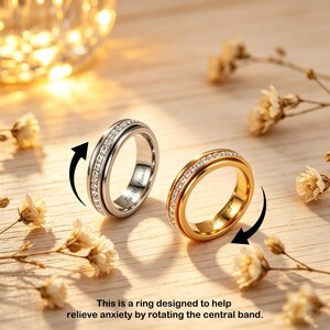 May include: Two rings, one silver and one gold, each with a rotating central band. Both rings are adorned with small, clear stones. The silver ring has the inscription "In My Heart". The text at the bottom says, "This is a ring designed to help relieve anxiety by rotating the central band."