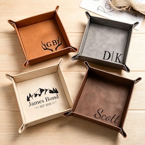 May include: Four square valet trays in tan, gray, cream, and dark brown. The tan tray features antlers and the letters GB. The gray tray shows D|K. The cream tray displays a mountain graphic with the text James Bond SET 2026. The dark brown tray features the name Scott.