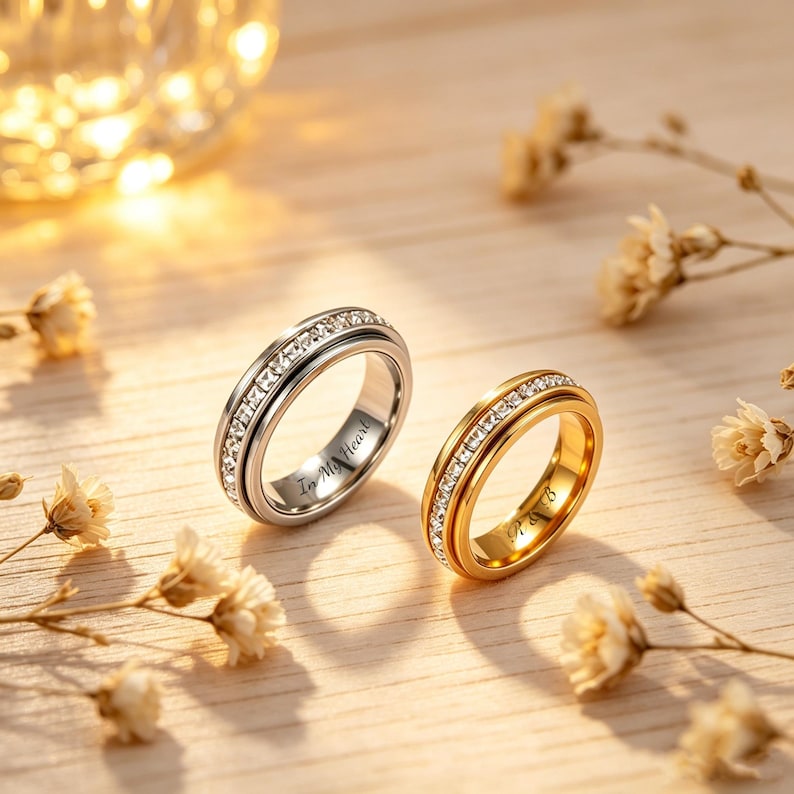 May include: Two rings, one silver and one gold, are displayed on a wooden surface. Both rings feature a band of clear stones. The silver ring has the inscription "In My Heart". The gold ring has the inscription "P & B".