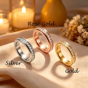May include: Three rings in silver, rose gold, and gold, each with a band of small, clear gemstones. The rings are displayed on a soft, neutral-toned surface. The words "Silver", "Rose Gold", and "Gold" are written on the image.