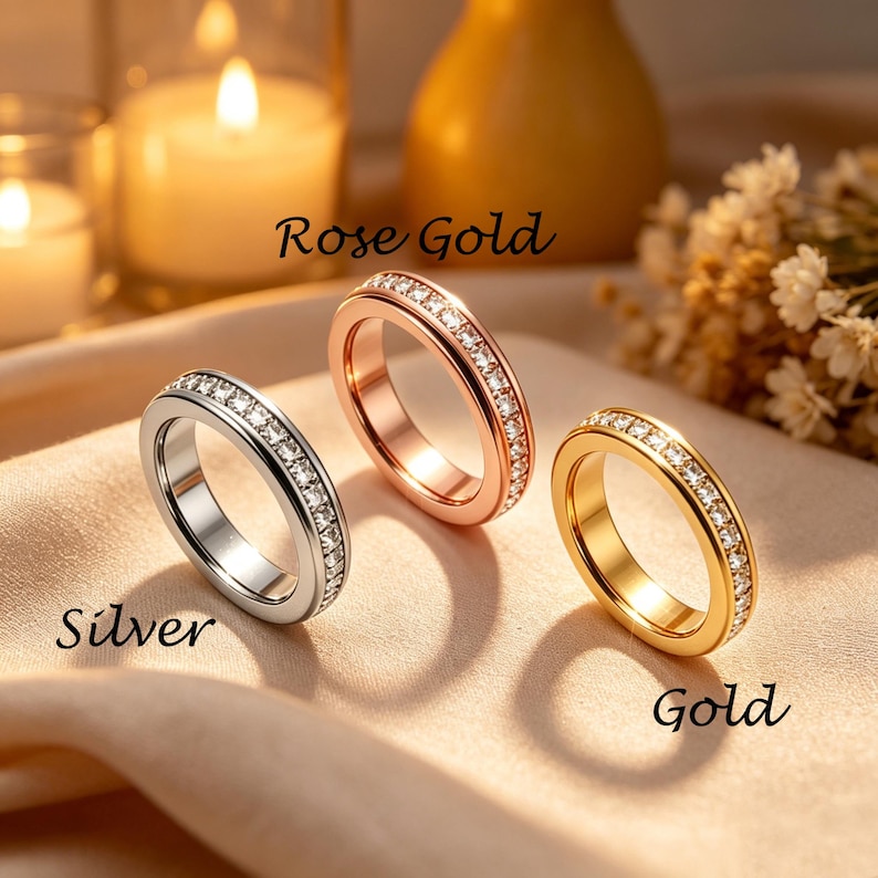 May include: Three rings in silver, rose gold, and gold, each with a band of small, clear gemstones. The rings are displayed on a soft, neutral-toned surface. The words "Silver", "Rose Gold", and "Gold" are written on the image.