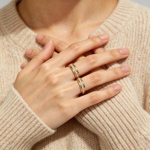 May include: Two gold rings with a row of small, clear gemstones. The rings are on a person's fingers, with the hands crossed over the chest. The rings have a simple, elegant design and are set against a beige sweater.