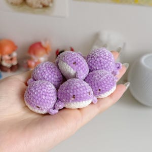 Puede incluir: Colección de pequeñas ballenas de ganchillo hechas a mano en tonos lavanda y blanco. Las ballenas tienen ojos pequeños y oscuros y se sostienen en una mano. Los peluches son suaves y redondos, con una superficie texturizada.