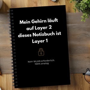 Könnte beinhalten: Ein schwarzes Notizbuch mit Spiralbindung und weißem Text: Mein Gehirn läuft auf Layer 2 dieses Notizbuch ist Layer 1. Unter dem Text befindet sich ein goldenes Vorhängeschloss-Symbol, gefolgt von: Kein WLAN erforderlich. 100% analog.
